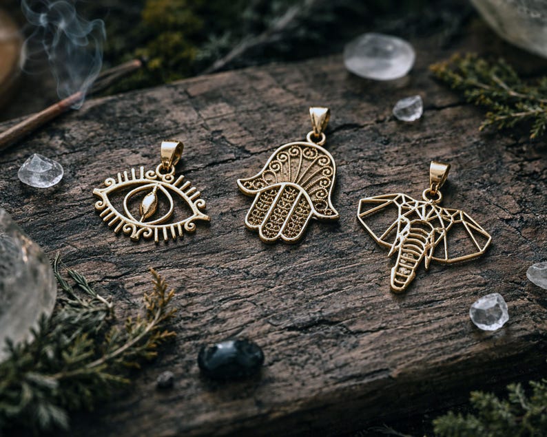 May include: Three gold-coloured pendants with detailed designs are displayed on a weathered wooden surface. The pendants feature an eye, a Hamsa hand, and an elephant. The background includes crystals, greenery, and a lit incense stick, creating a mystical aesthetic.