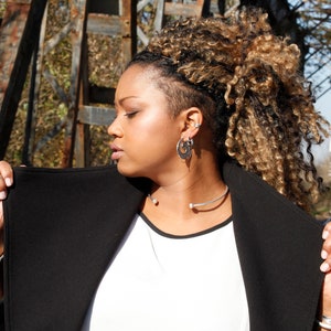 May include: A woman with long, blonde hair is wearing a black jacket over a white shirt. She is wearing a silver necklace and earrings.