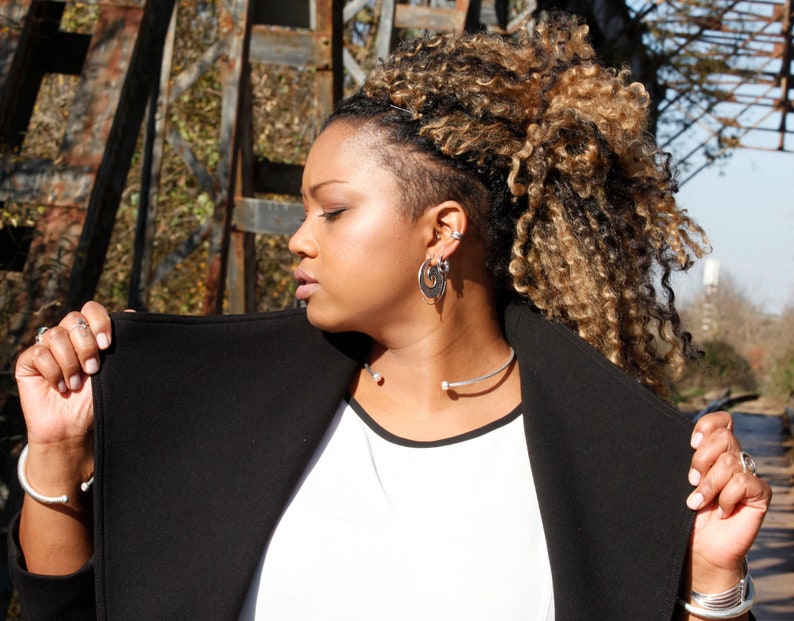 May include: A woman with long, blonde hair is wearing a black jacket over a white shirt. She is wearing a silver necklace and earrings.