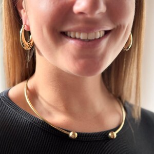 May include: A gold choker necklace with two round balls at the ends. The necklace is worn by a person with long blonde hair and is sitting on their neck.