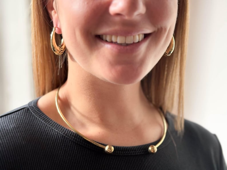 May include: A gold choker necklace with two round balls at the ends. The necklace is worn by a person with long blonde hair and is sitting on their neck.