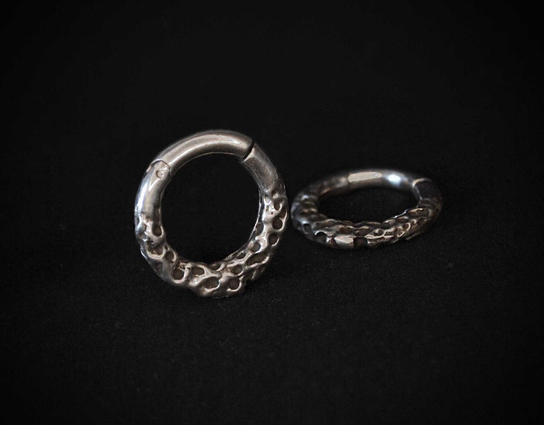 May include: Two silver-toned circular hoop rings featuring a textured, pitted surface on the outer edge. Each ring has a smooth, plain segment near the hinge mechanism, contrasting with the irregular, crater-like detailing on the rest of the band.