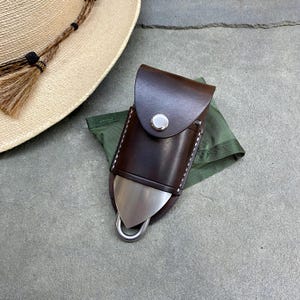 May include: A dark brown leather sheath with white stitching and a silver-coloured metal snap closure. The sheath contains a metallic tool with a pointed, brushed-finish blade and a rounded silver loop at the bottom, resting on a folded olive green fabric.