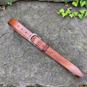 May include: A brown leather belt with a silver buckle. The belt is lying on a gray, textured surface.