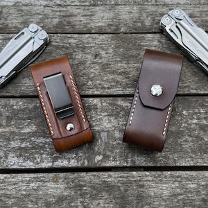 May include: Two multi-tools made of silver-toned metal are positioned next to two leather sheaths. One sheath is light brown with a black metal belt clip, and the other is dark brown with a silver-toned snap button closure and white stitching.