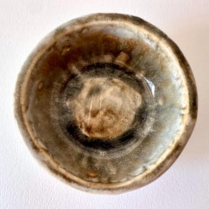 May include: A small, round ceramic bowl featuring a reactive glaze with blended tones of earthy brown, charcoal grey, and creamy white. The interior surface displays a smooth, glossy finish with organic colour variations and a thick, rounded rim.