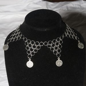 May include: A silver-toned necklace with a geometric design. The necklace features interconnected circles and dangling coin-shaped pendants. The necklace is displayed on a black mannequin head against a white background.
