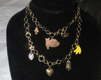 May include: Two gold-coloured layered necklaces with assorted charms, including stars, hearts, a cat, and a banana. The necklaces are displayed on a black mannequin. The charms are in various colours, including gold, brown, yellow, and blue.