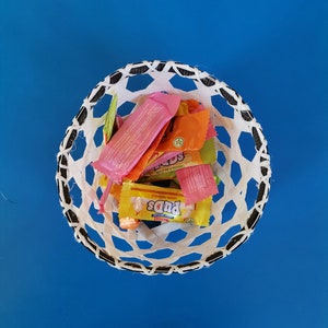 May include: A white, open-weave basket with a black trim, filled with colourful candy wrappers. The wrappers are pink, orange, yellow, and green. The basket sits on a bright blue background.