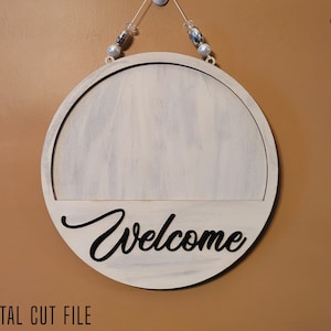 May include: A white wooden round sign with the word "Welcome" in black script lettering. The sign is hanging from a white hook with a string of beads.