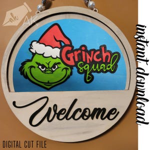 May include: A round wooden sign with a white background and a blue circle in the center. The circle features a green Grinch character with a red hat and the text "Grinch Squad". Below the circle is the text "Welcome" in black.