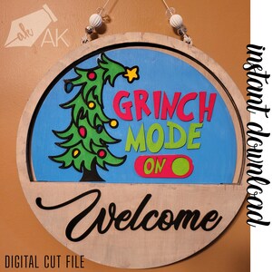 May include: A round wooden sign with a blue background and a green Christmas tree with colorful ornaments. The text "Grinch Mode On" is written in red, green, and pink letters. The bottom of the sign is wood with the word "Welcome" written in black.