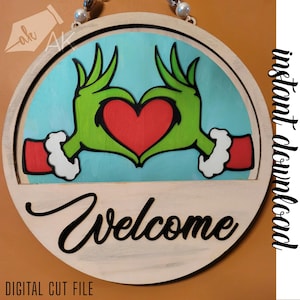 May include: A round wooden sign with a green Grinch hand holding a red heart. The sign says "Welcome" in black lettering.