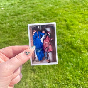 May include: A small, rectangular sticker depicting a historical painting of three figures in a doorway. One figure wears a long blue dress, another a red and pink outfit, and the third a dark outfit. The sticker is held against a backdrop of green grass.