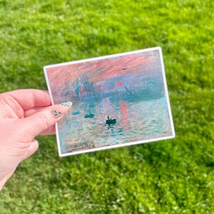 Op de afbeelding: Een sticker met een schilderij van een zonsopgang boven een wateroppervlak met boten en een rode zon aan de hemel. Het schilderij is in een gedempte kleurenpalet van blauw, groen en roze.