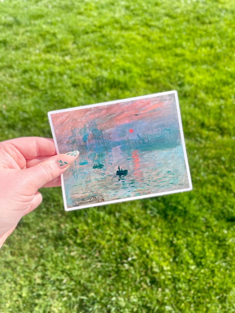 Op de afbeelding: Een sticker met een schilderij van een zonsopgang boven een wateroppervlak met boten en een rode zon aan de hemel. Het schilderij is in een gedempte kleurenpalet van blauw, groen en roze.