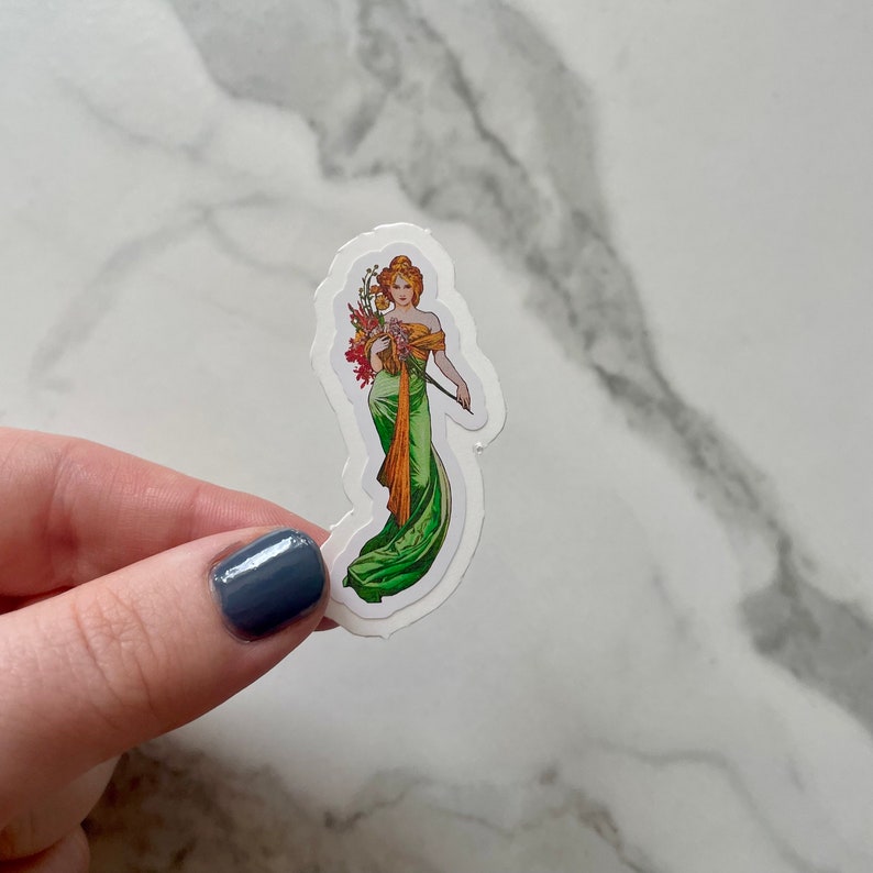 May include: A sticker featuring a woman in a green dress with a floral bouquet. She is holding a staff and has a crown of flowers on her head.