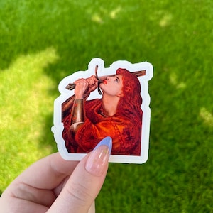 May include: A sticker featuring a painting of a woman with red hair holding a sword above her head. The woman is wearing a red and gold robe and is looking up at the sword.