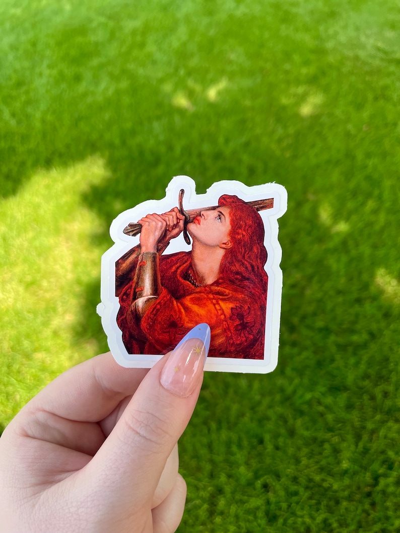 May include: A sticker featuring a painting of a woman with red hair holding a sword above her head. The woman is wearing a red and gold robe and is looking up at the sword.