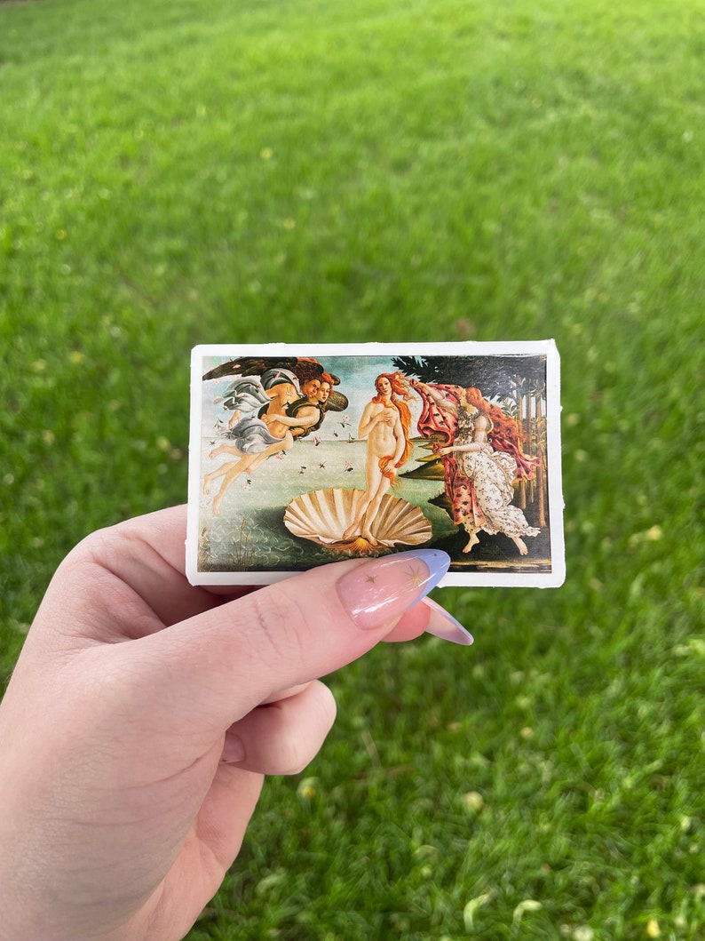 May include: A sticker featuring a reproduction of Sandro Botticelli's The Birth of Venus painting. The painting depicts a nude woman standing on a giant scallop shell, with the god of the wind, Zephyrus, and the nymph, Chloris, blowing her towards the shore.