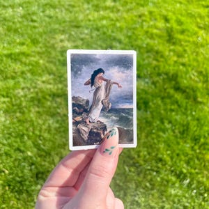 May include: A sticker featuring a painting of a woman in a white dress standing on a rocky cliff, looking out at a stormy sea.