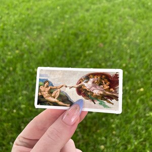 May include: A close-up of a hand holding a sticker featuring a reproduction of Michelangelo's The Creation of Adam painting. The sticker is white with a blue border and shows God reaching out to Adam.