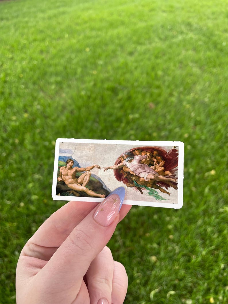 May include: A close-up of a hand holding a sticker featuring a reproduction of Michelangelo's The Creation of Adam painting. The sticker is white with a blue border and shows God reaching out to Adam.