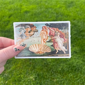 May include: A vintage postcard featuring a painting of the Birth of Venus by Sandro Botticelli. The painting depicts a nude woman standing on a giant scallop shell, with the wind blowing her hair and a flowing drapery behind her. Two winged figures are shown in the background, one holding a red cloak and the other holding a flower.