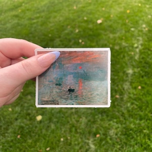 Op de afbeelding: Een sticker met een schilderij van een zonsopgang boven een wateroppervlak met twee kleine boten. De lucht is een mix van oranje, roze en blauw met een rode zon.