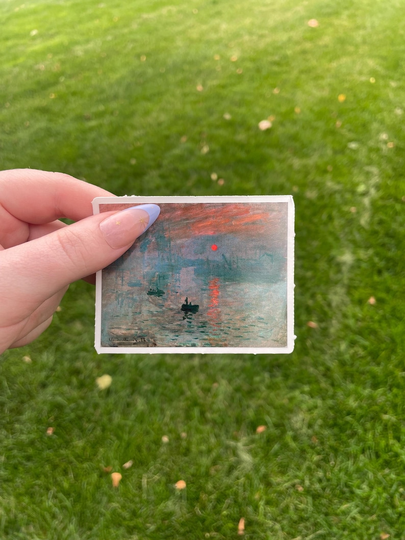 Op de afbeelding: Een sticker met een schilderij van een zonsopgang boven een wateroppervlak met twee kleine boten. De lucht is een mix van oranje, roze en blauw met een rode zon.