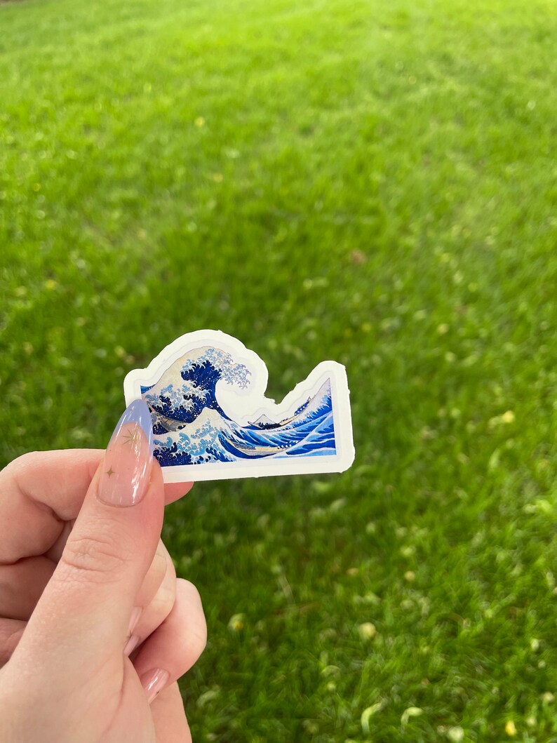 Może przedstawiać: Naklejka przedstawiająca niebiesko-białą wersję "Wielkiej Fali w Kanagawie" autorstwa Katsushiki Hokusai. Naklejka jest trzymana w dłoni z niebieskim i białym lakierem do paznokci.