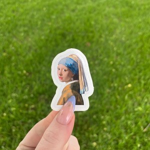 May include: A white sticker with a portrait of a woman wearing a blue and yellow headscarf and a pearl earring. The woman is looking to the side and has a light complexion.