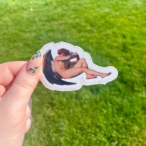 Puede incluir: Una pegatina con una pintura de una figura alada con cabello rojo, sentada con los brazos cruzados y mirando hacia abajo. La figura está desnuda y tiene un ala grande y negra.