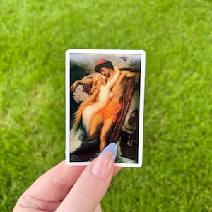 May include: A sticker featuring a painting of a couple embracing. The man is wearing a red hat and the woman is wearing a white dress. The painting is set against a dark blue background with waves and a boat.