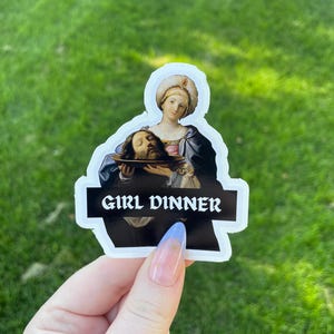 Op de afbeelding: Een witte sticker met een zwarte achtergrond waarop een schilderij te zien is van een vrouw die een bord vasthoudt met daarop een mannenhoofd. De tekst "GIRL DINNER" is in wit gedrukt op een zwarte banner onder het schilderij.