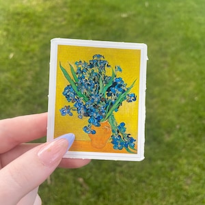 May include: A sticker featuring a painting of a bouquet of blue irises in a yellow vase on a yellow background.