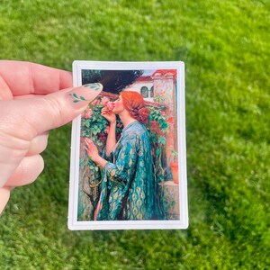 May include: A woman in a blue and green patterned robe is smelling a pink rose. She is standing in a garden with a white building in the background.