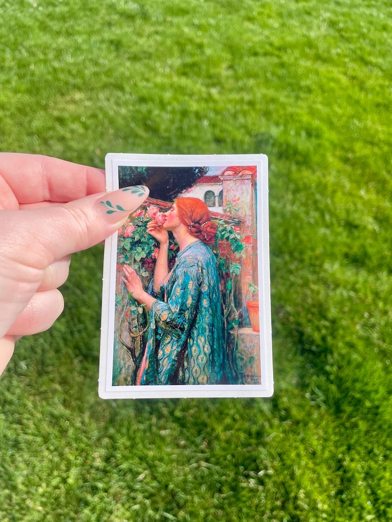 May include: A woman in a blue and green patterned robe is smelling a pink rose. She is standing in a garden with a white building in the background.