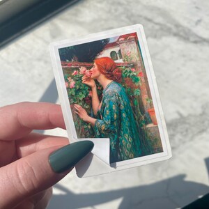 May include: A painting of a woman with red hair smelling a pink rose. She is wearing a blue and green robe with gold accents. The background is a garden with green leaves and pink roses.