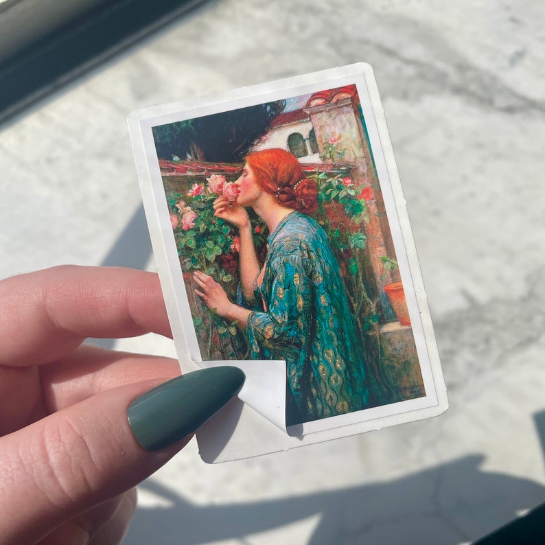 May include: A painting of a woman with red hair smelling a pink rose. She is wearing a blue and green robe with gold accents. The background is a garden with green leaves and pink roses.