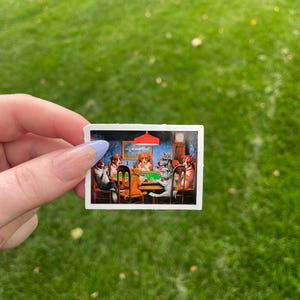 May include: A sticker featuring a painting of dogs playing cards around a green table. The dogs are wearing clothes and have human-like expressions. The painting is set in a room with a red lampshade and a clock on the wall.