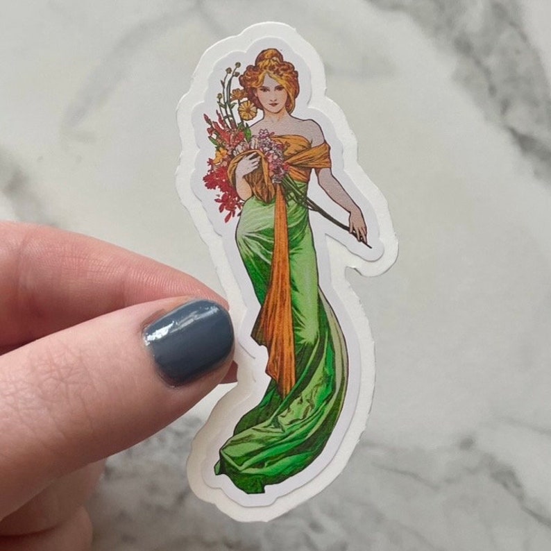 May include: A sticker featuring a vintage illustration of a woman in a green gown holding a bouquet of flowers. The woman has long brown hair and is wearing a gold sash.