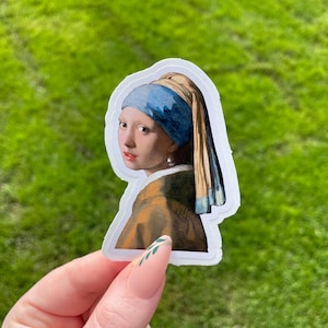 Pode incluir: Um adesivo com uma pintura de uma mulher com um lenço azul e branco e um brinco de pérola. O adesivo está sobre um fundo verde gramado.