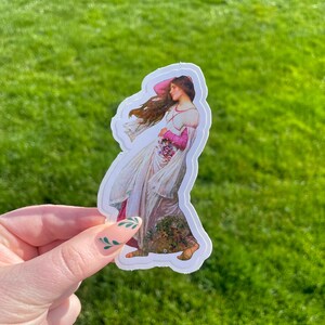 May include: A sticker featuring a painting of a woman in a white dress with pink accents, holding a bouquet of flowers. The woman is standing in a field of flowers.