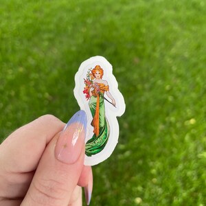 May include: A white sticker with a vintage illustration of a woman in a green dress holding a bouquet of flowers. The woman has red hair and is wearing a crown.