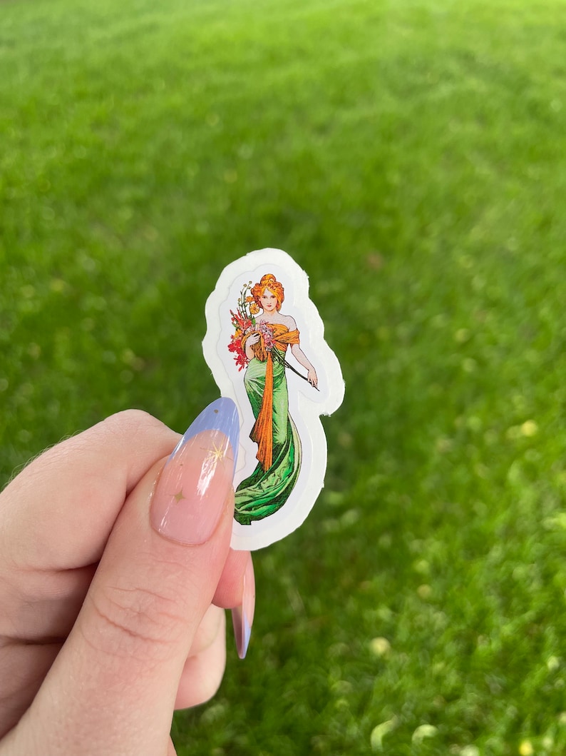 May include: A white sticker with a vintage illustration of a woman in a green dress holding a bouquet of flowers. The woman has red hair and is wearing a crown.