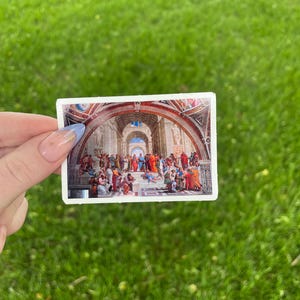 May include: A sticker featuring a painting of a group of people in a large room with a vaulted ceiling. The people are dressed in colorful robes and appear to be engaged in a discussion.