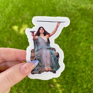 May include: A sticker featuring a vintage illustration of a woman in a flowing blue gown, holding a goblet and a staff, seated on a throne with lion head armrests.