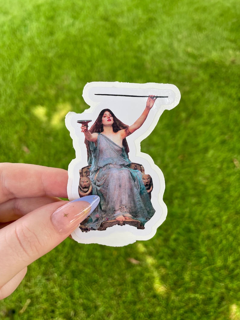 May include: A sticker featuring a vintage illustration of a woman in a flowing blue gown, holding a goblet and a staff, seated on a throne with lion head armrests.