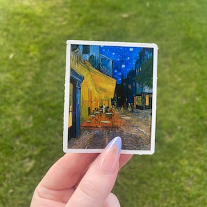 May include: A sticker featuring a painting of a cafe at night with a starry sky. The cafe is lit with yellow lights and there are people sitting at tables outside. The painting is in a style reminiscent of Vincent van Gogh.